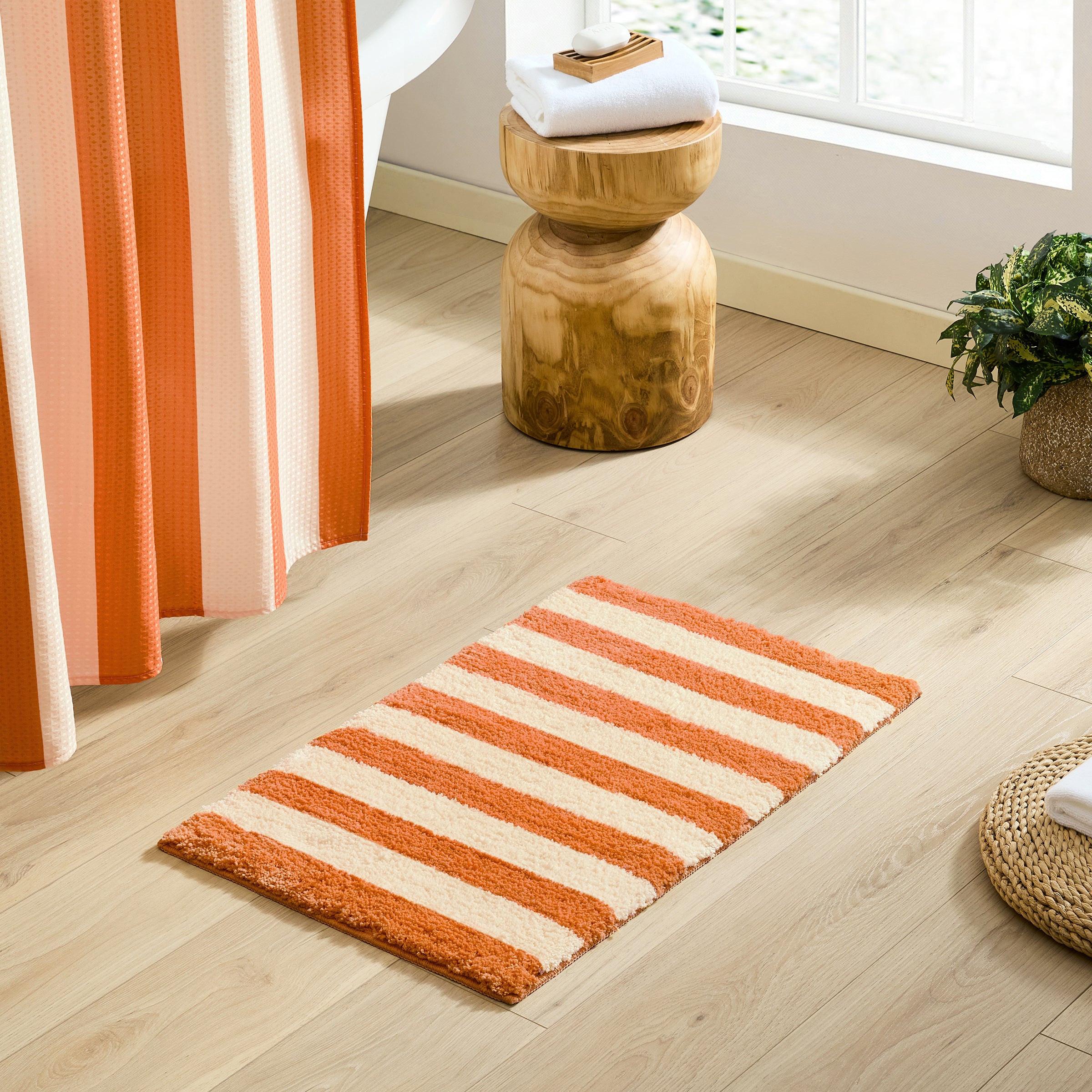 A rectangular bath mat with alternating orange and cream stripes lies on a light wood bathroom floor. A matching orange and cream shower curtain hangs on the left, with a small wooden stool, folded white towels, and a potted plant near a bright window in the background.