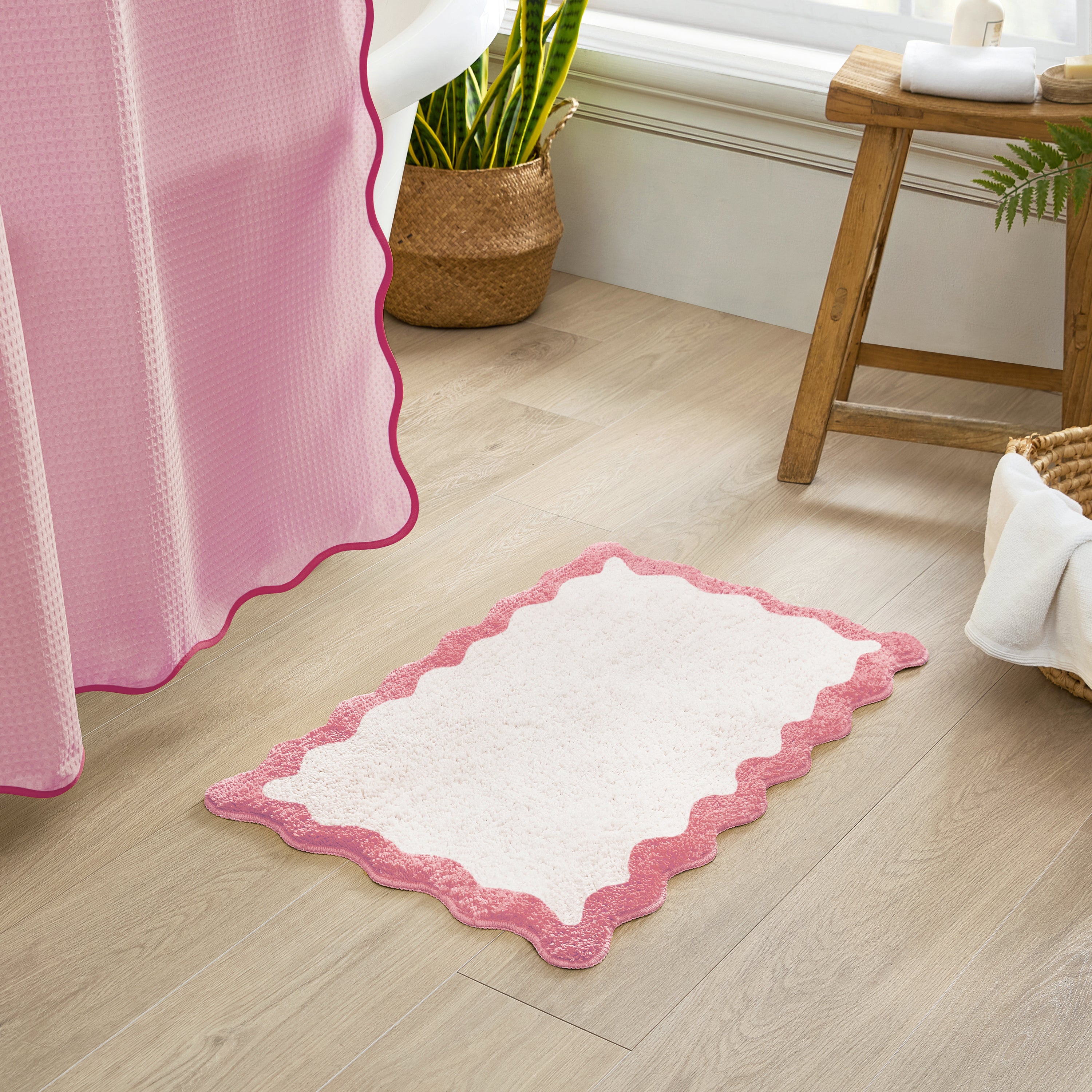 White bath mat with pink border on a wooden floor next to a pink shower curtain.