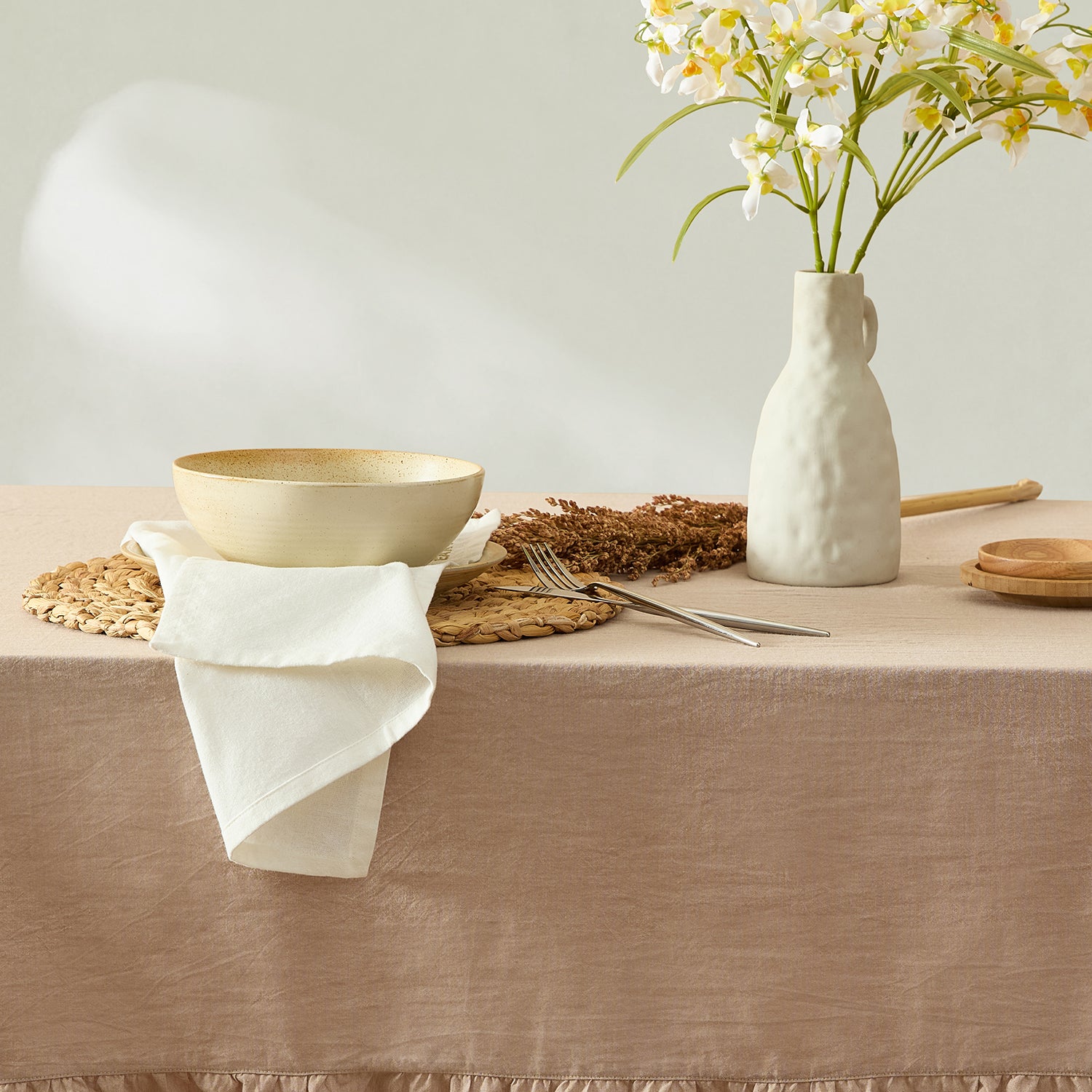 Table setting with a beige bowl, white vase with flowers, and cutlery on a beige tablecloth.