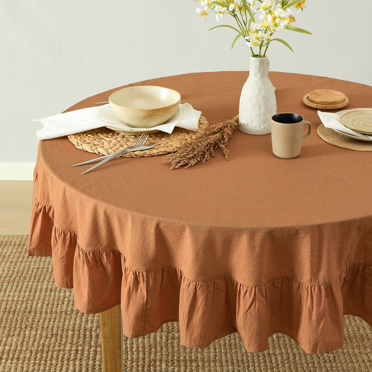 Round table with a brown ruffled tablecloth, plates, cups, and a vase with flowers.