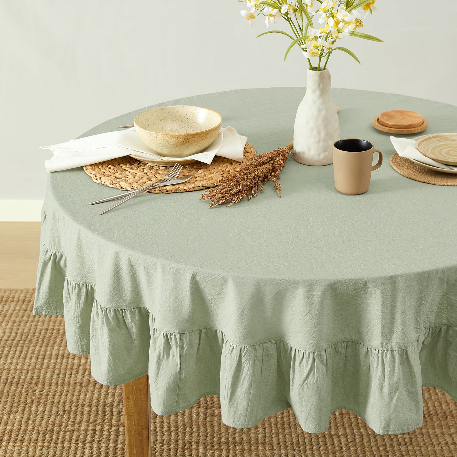 Round table with a green tablecloth, plates, cups, and a vase with flowers on a light background.