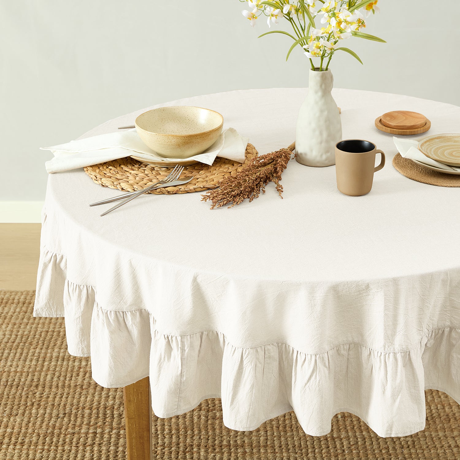Round table with a white tablecloth, plates, bowls, and a vase with flowers on a neutral background.