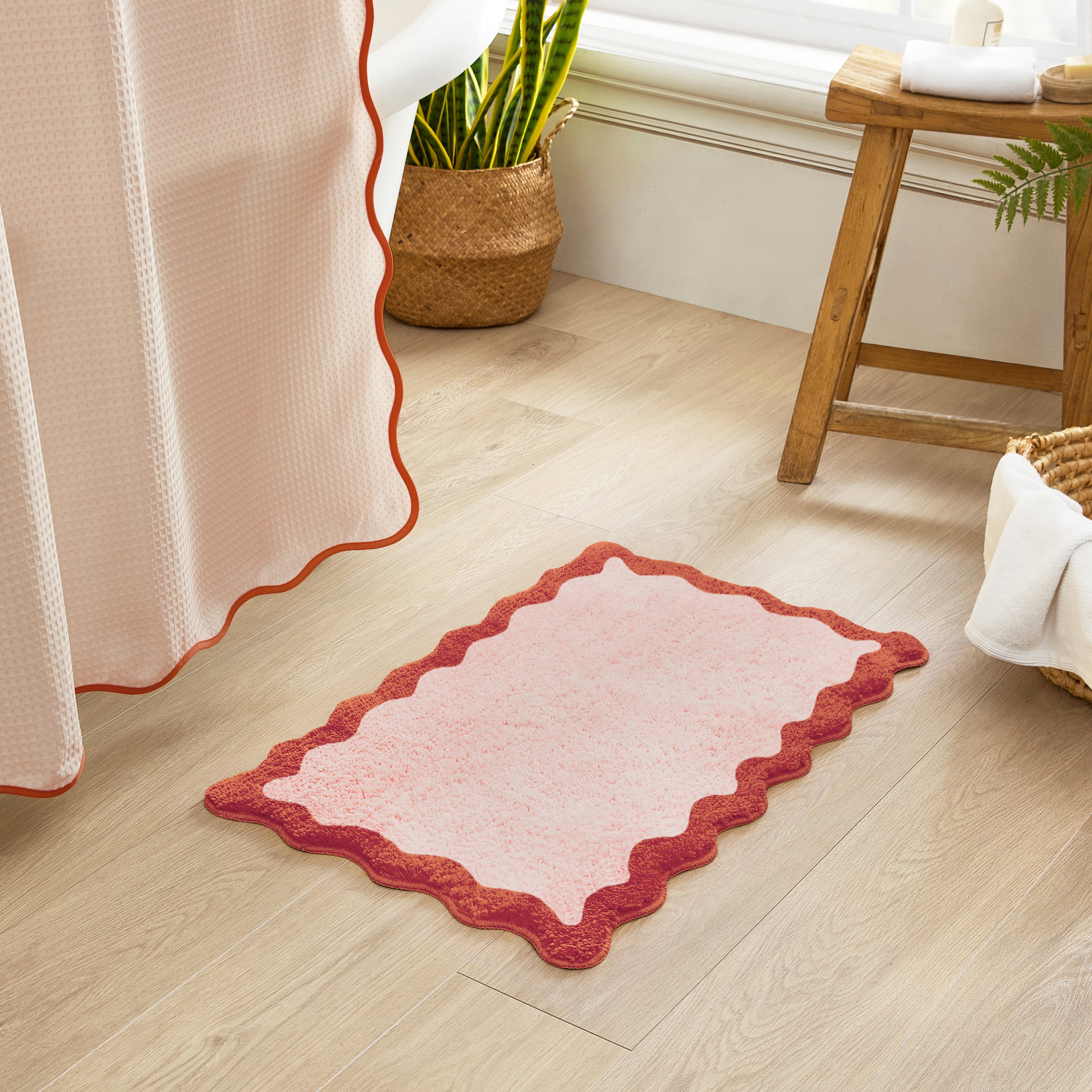 Pink bath mat with red border on a wooden floor next to a curtain and plant.