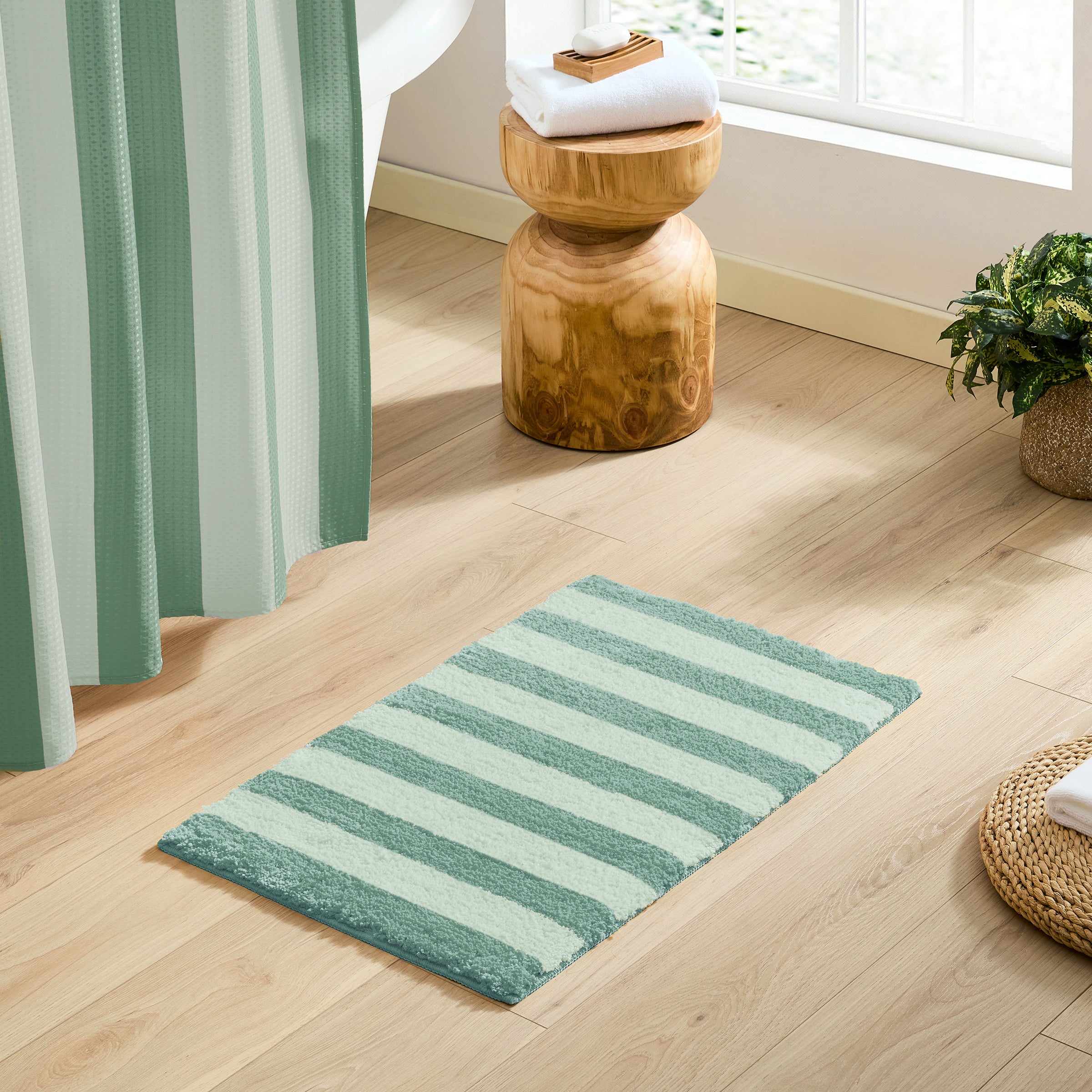 A rectangular bath mat with horizontal stripes in sage green and white, styled on a light wood floor in a bright bathroom setting.