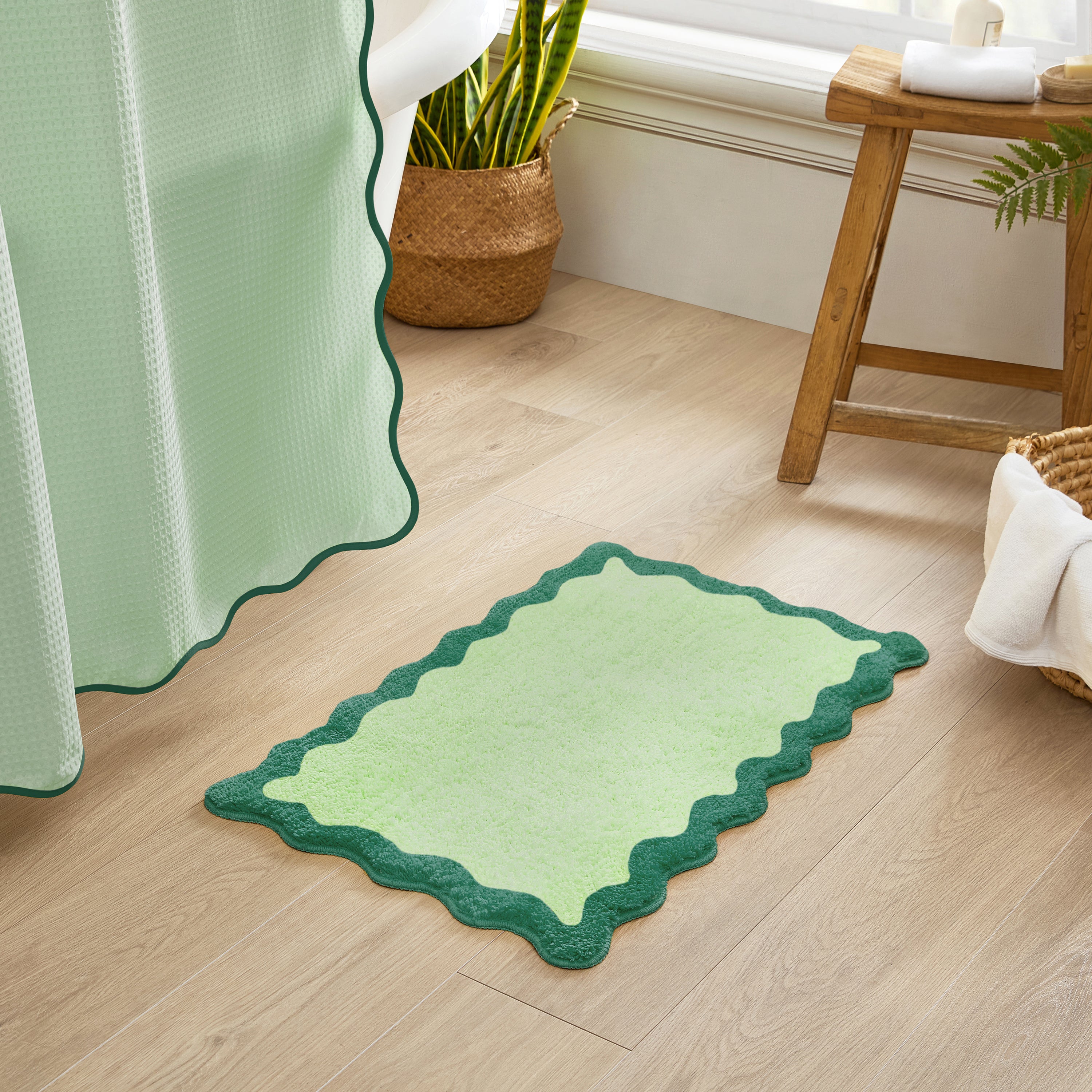Green bath mat with scalloped edge on a wooden floor, next to a window with a plant and a wooden stool.