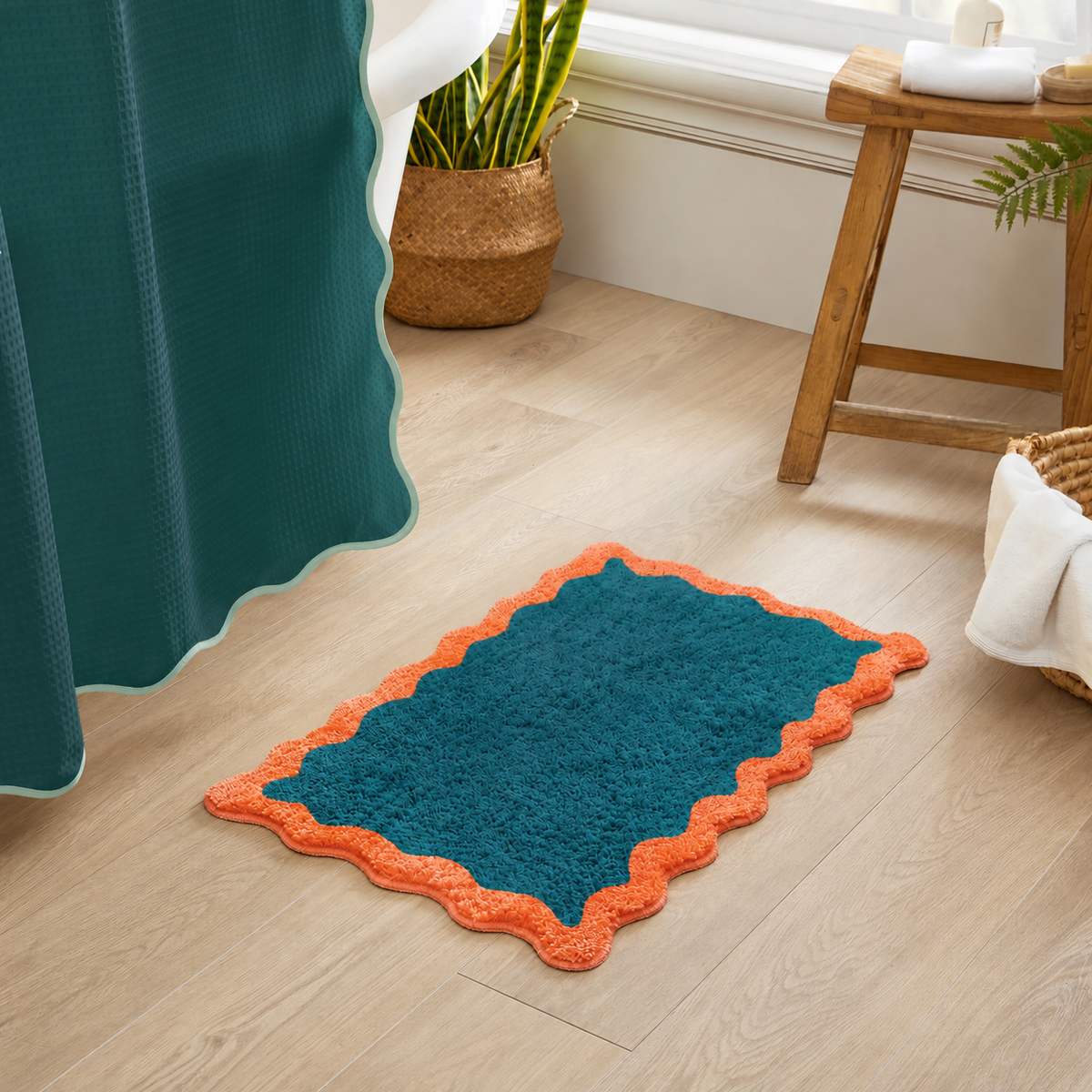 A rectangular bath mat in teal with an orange scalloped border, placed on a light wood floor in a bathroom setting next to a matching teal shower curtain.