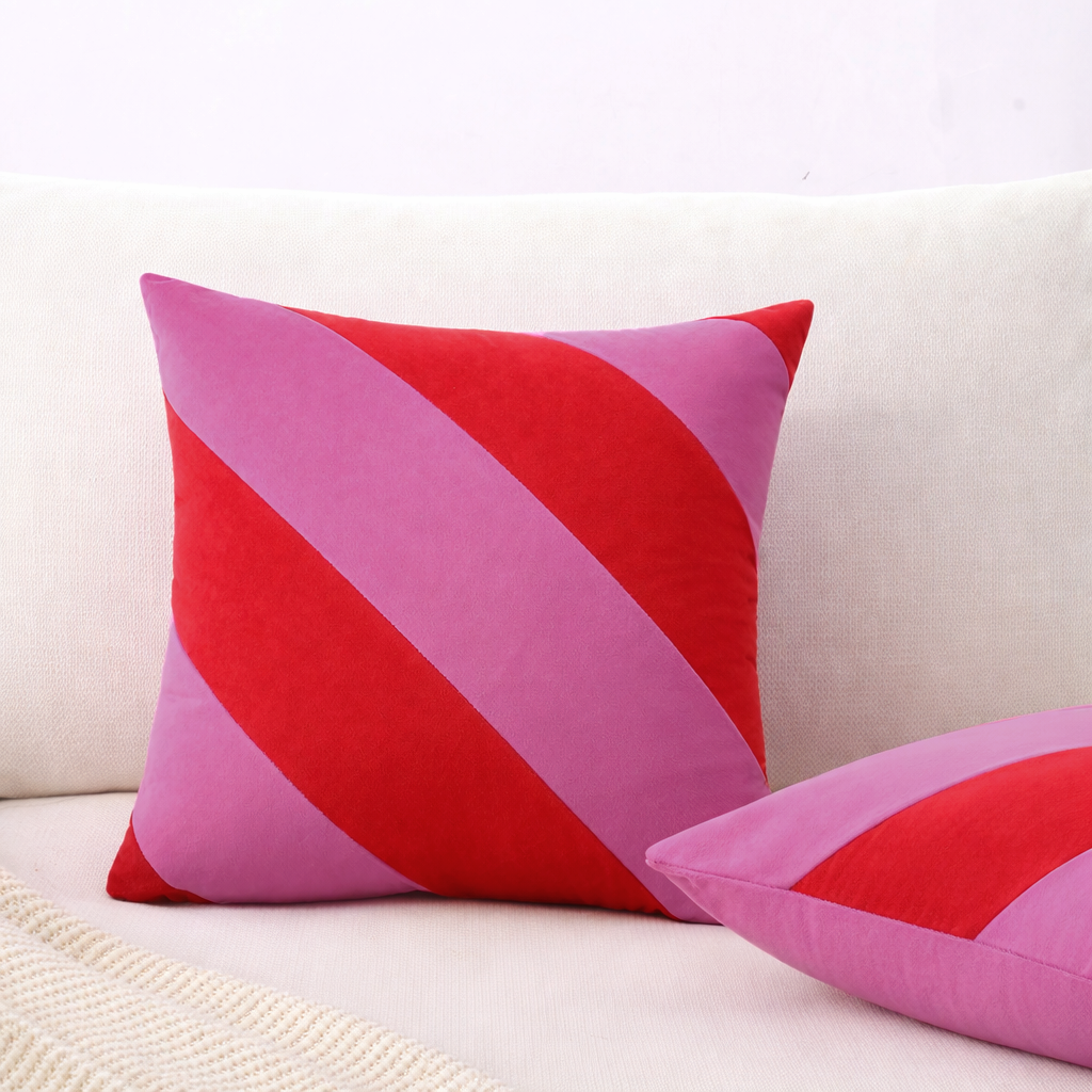 A square throw pillow with diagonal stripes in red, pink, and purple, styled on a light beige sofa next to another matching pillow.