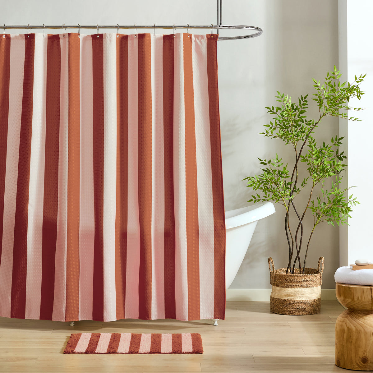 A striped shower curtain hanging in a bathroom, with vertical bands in rust red, terracotta, blush pink, and cream. A matching bath mat is on the floor, with a white tub, potted plant, and wooden stool nearby.