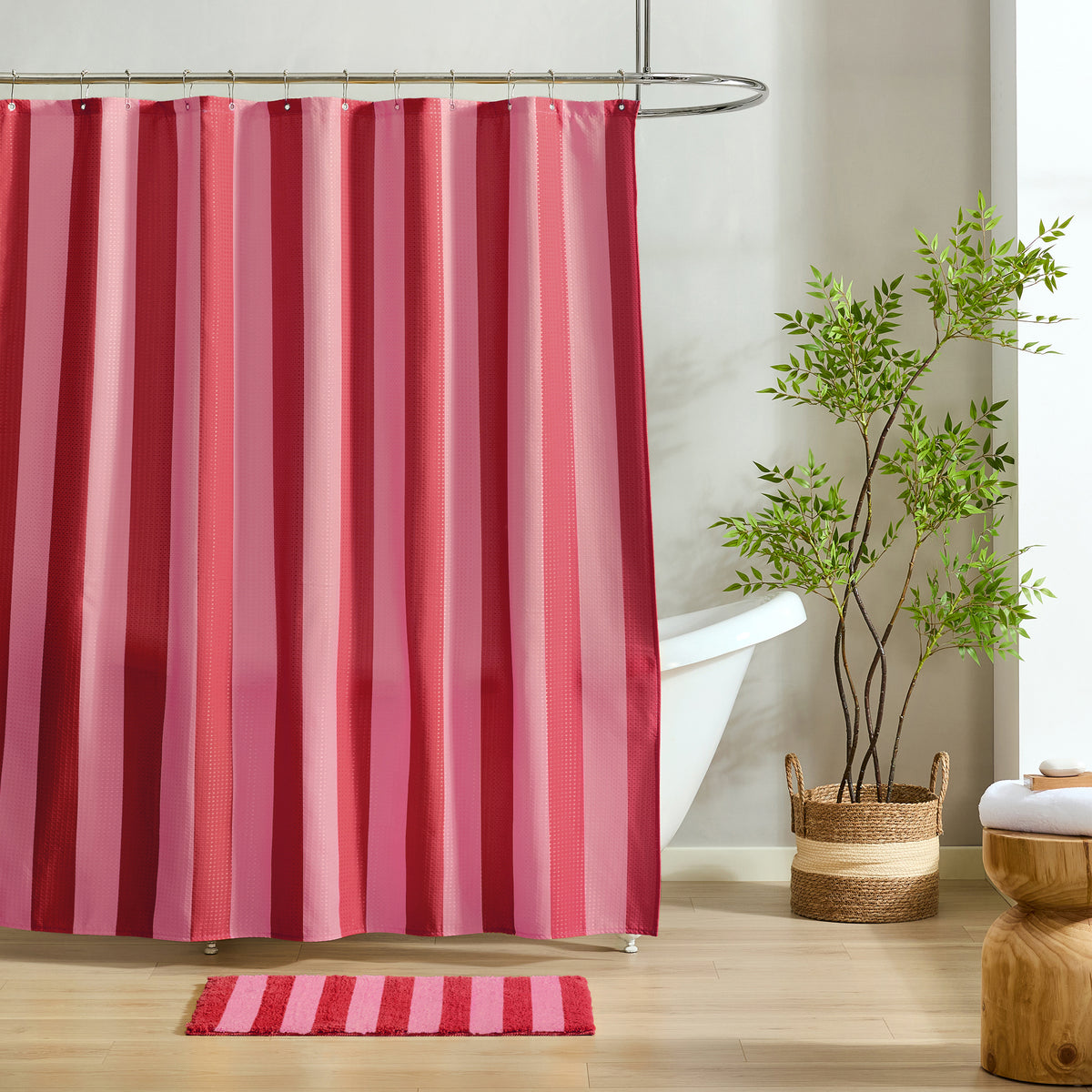 A pink and red striped shower curtain hanging in a bathroom setting with a white tub, a potted green plant, and a striped bath mat on the floor.