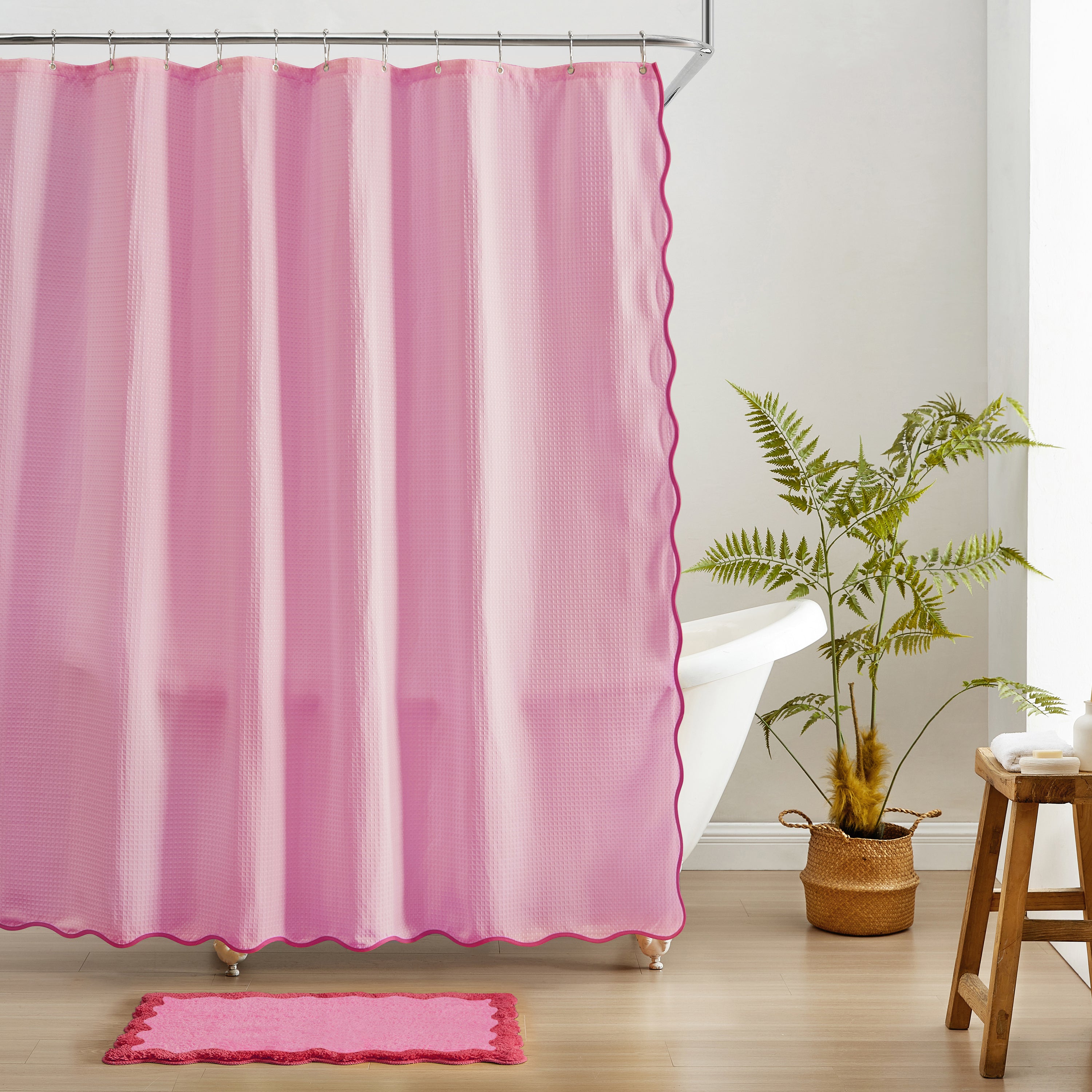 Pink shower curtain with scalloped edge in a bathroom setting with a plant and stool.