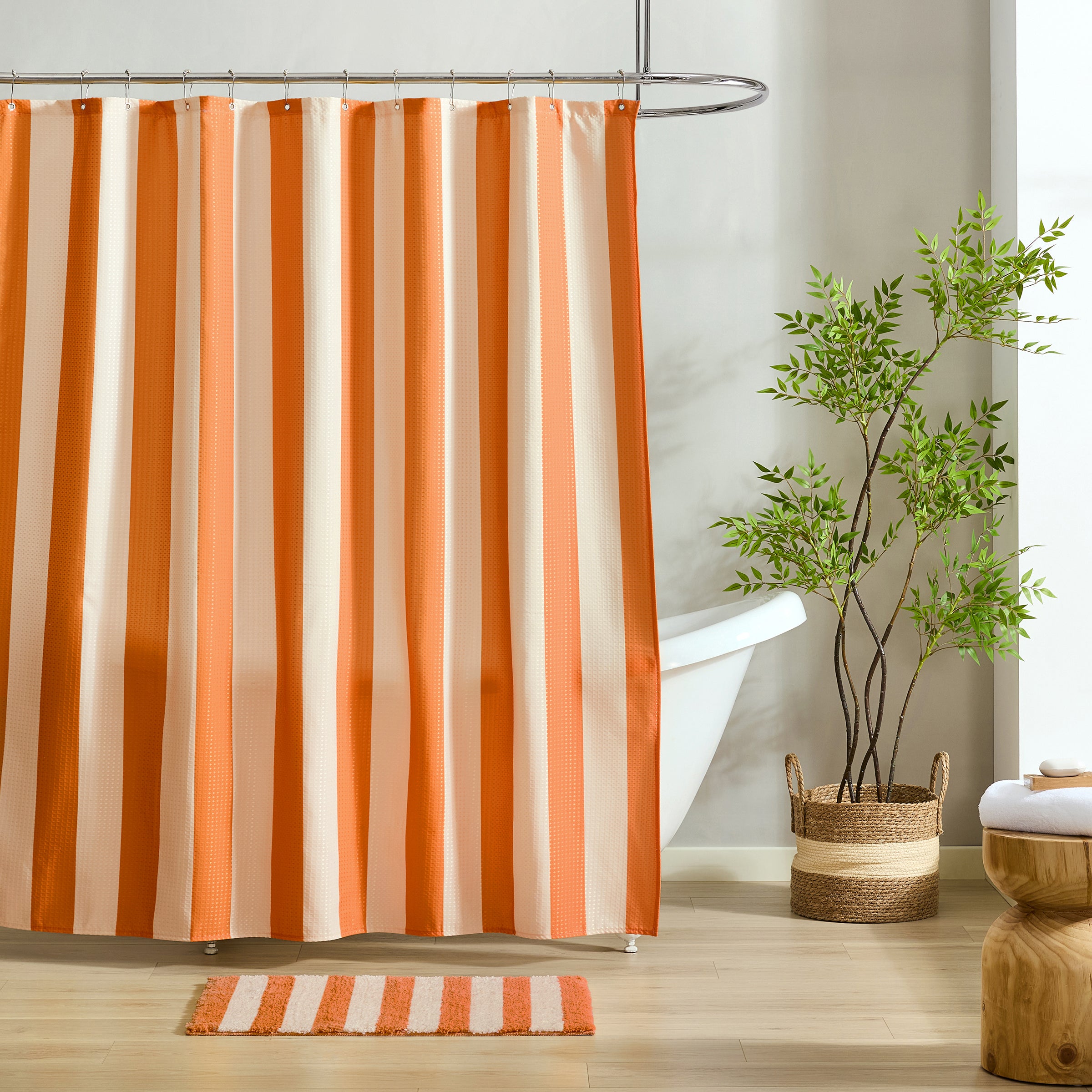An orange and cream striped shower curtain hanging in a bright bathroom with a white tub, a potted green plant, and a matching striped bath mat.