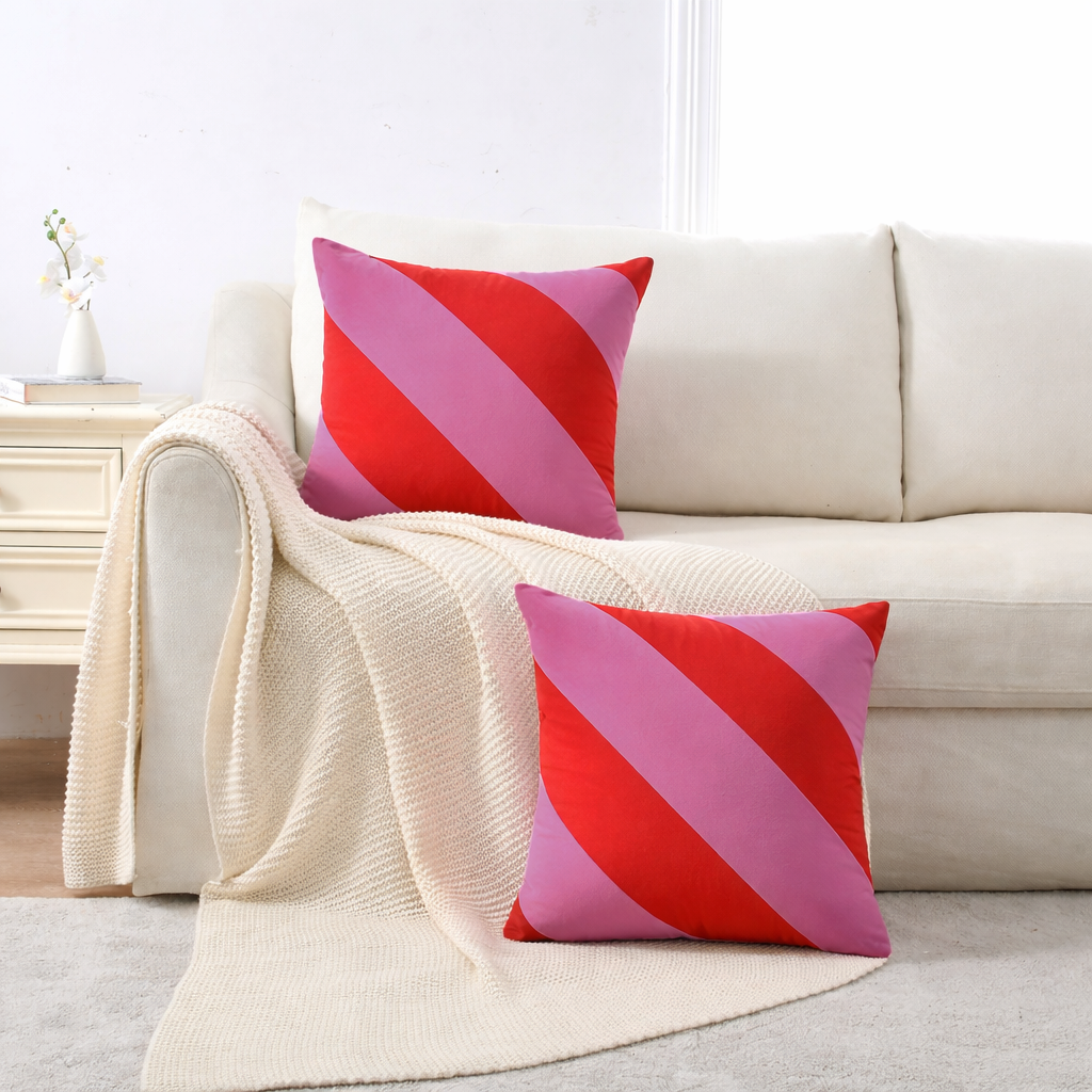 Two square throw pillows with diagonal pink and red stripes are styled on a cream sofa with a matching knit throw blanket in a bright living room setting.