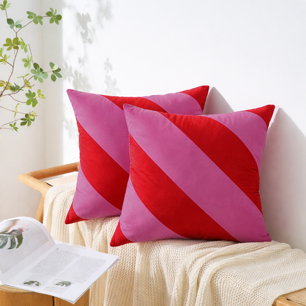 Two square throw pillows with diagonal stripes in red and pink, styled on a cream knit blanket on a wooden bench with a book and plant in a bright room.